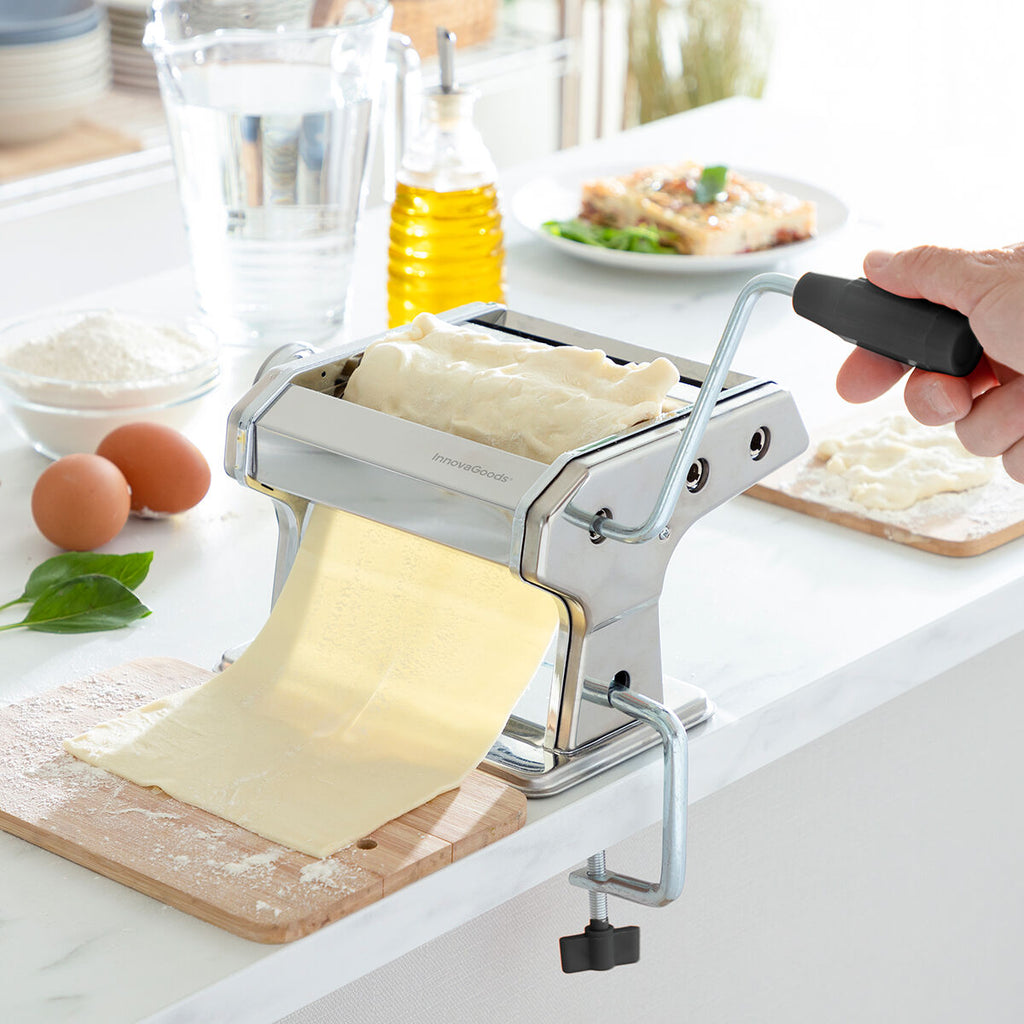 A pasta maker being used to roll out a sheet of pasta dough. The stainless steel device clamped to a kitchen counter. Visible ingredients include eggs, flour, and olive oil on a bright kitchen counter.
