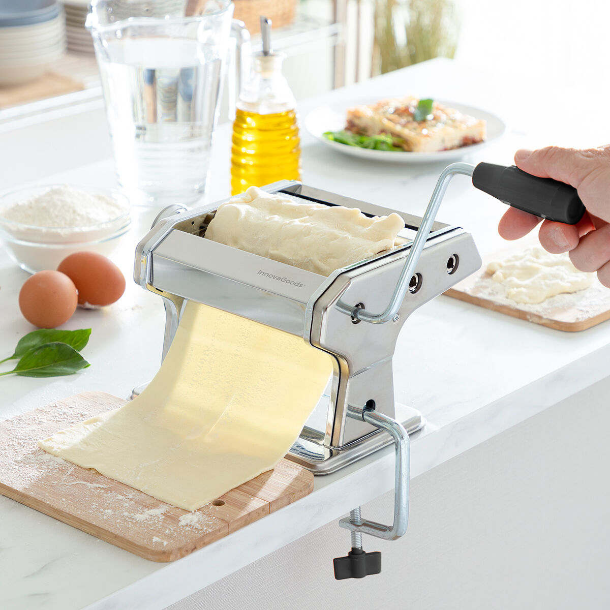 A pasta maker being used to roll out a sheet of pasta dough. The stainless steel device clamped to a kitchen counter. Visible ingredients include eggs, flour, and olive oil on a bright kitchen counter.