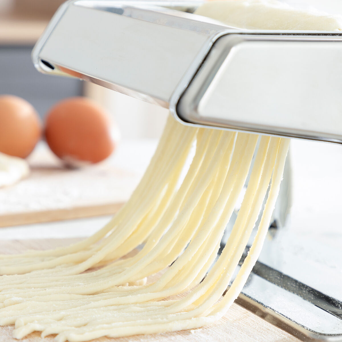 Close-up view of fresh spaghetti noodles emerging from the pasta maker, with eggs in the background.