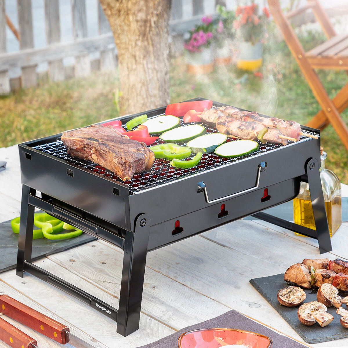 The portable grill is seen in a backyard setting with vegetables and meat cooking on top. The grill is black with foldable legs and has the InnovaGoods logo on the side.