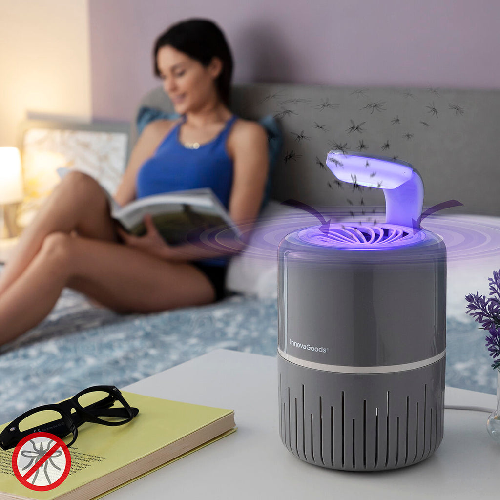 A lifestyle image showing the mosquito lamp on a bedside table with a woman reading in the background. The lamp is actively attracting insects, depicted by graphic representations of mosquitoes flying towards it. The setting is a bedroom with a cozy ambiance, and there is an emphasis on the lamp's effectiveness.