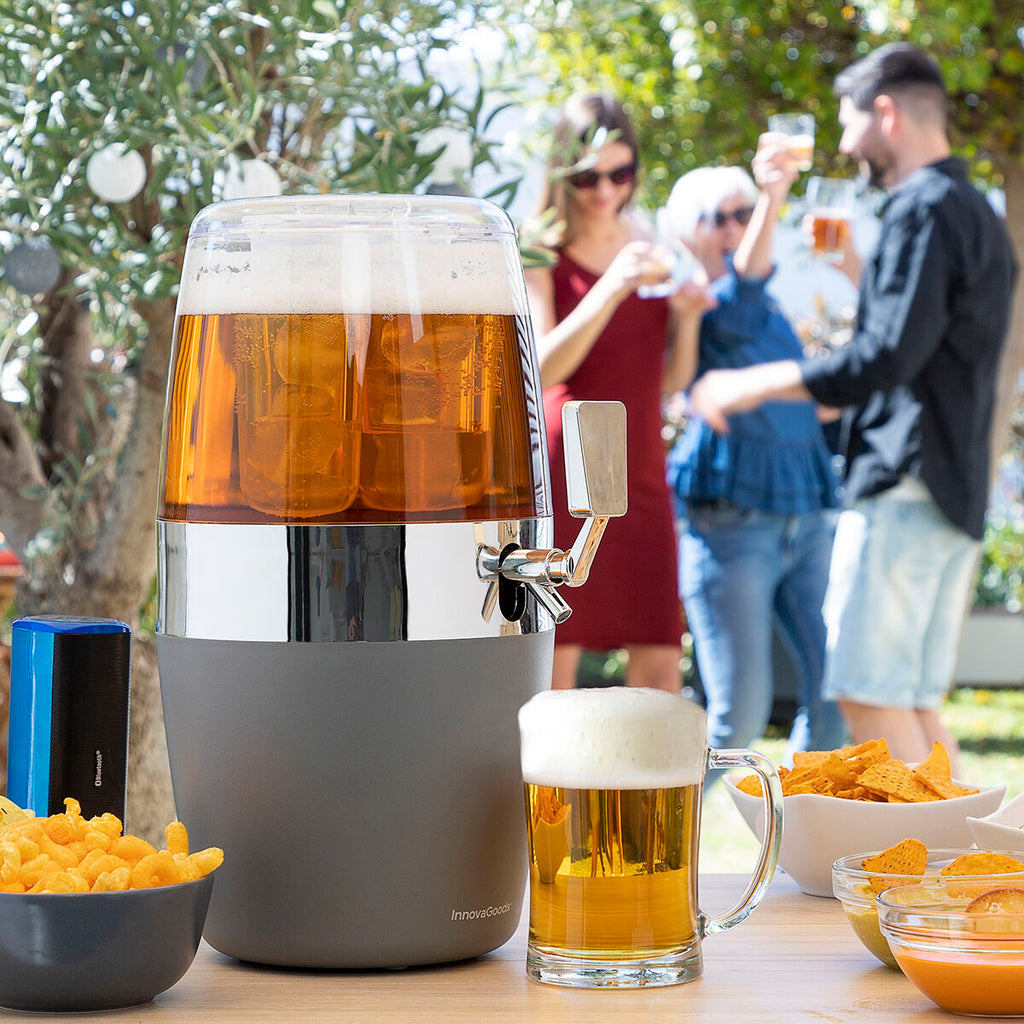 The cooling drinks dispenser in an outdoor party setting with people in the background enjoying their drinks. The dispenser is filled with a light beverage, possibly beer, ice is visible inside, and a filled mug sits beside it, surrounded by snacks.