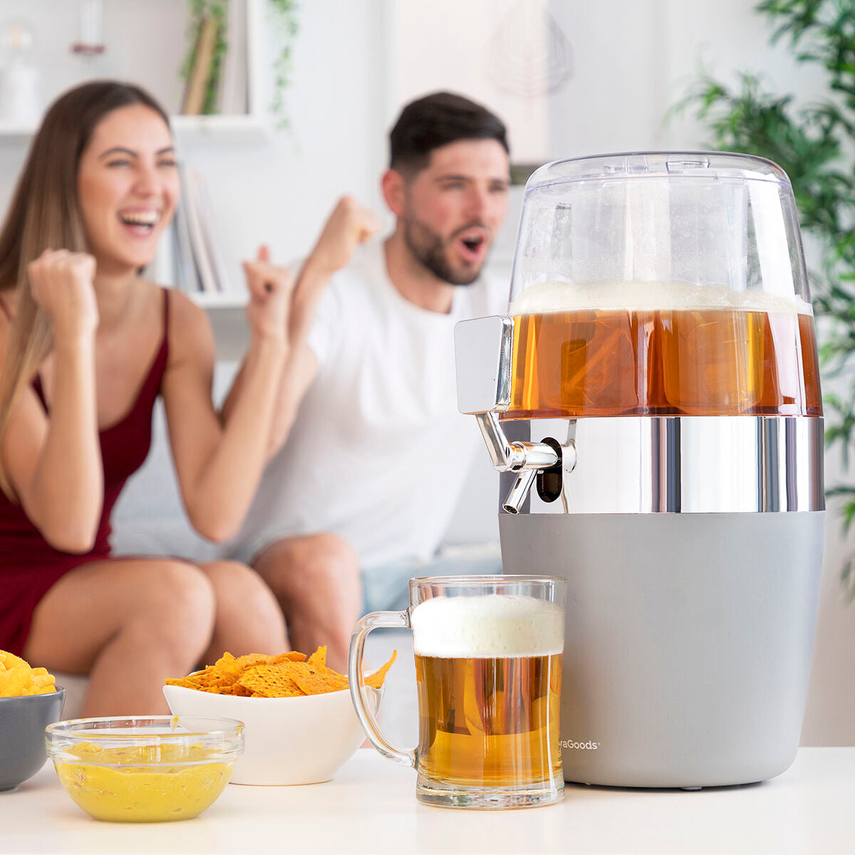 Indoor setting showing the dispenser filled with beer beside snacks, indicating a home entertainment scenario. People in the background appear to be celebrating.