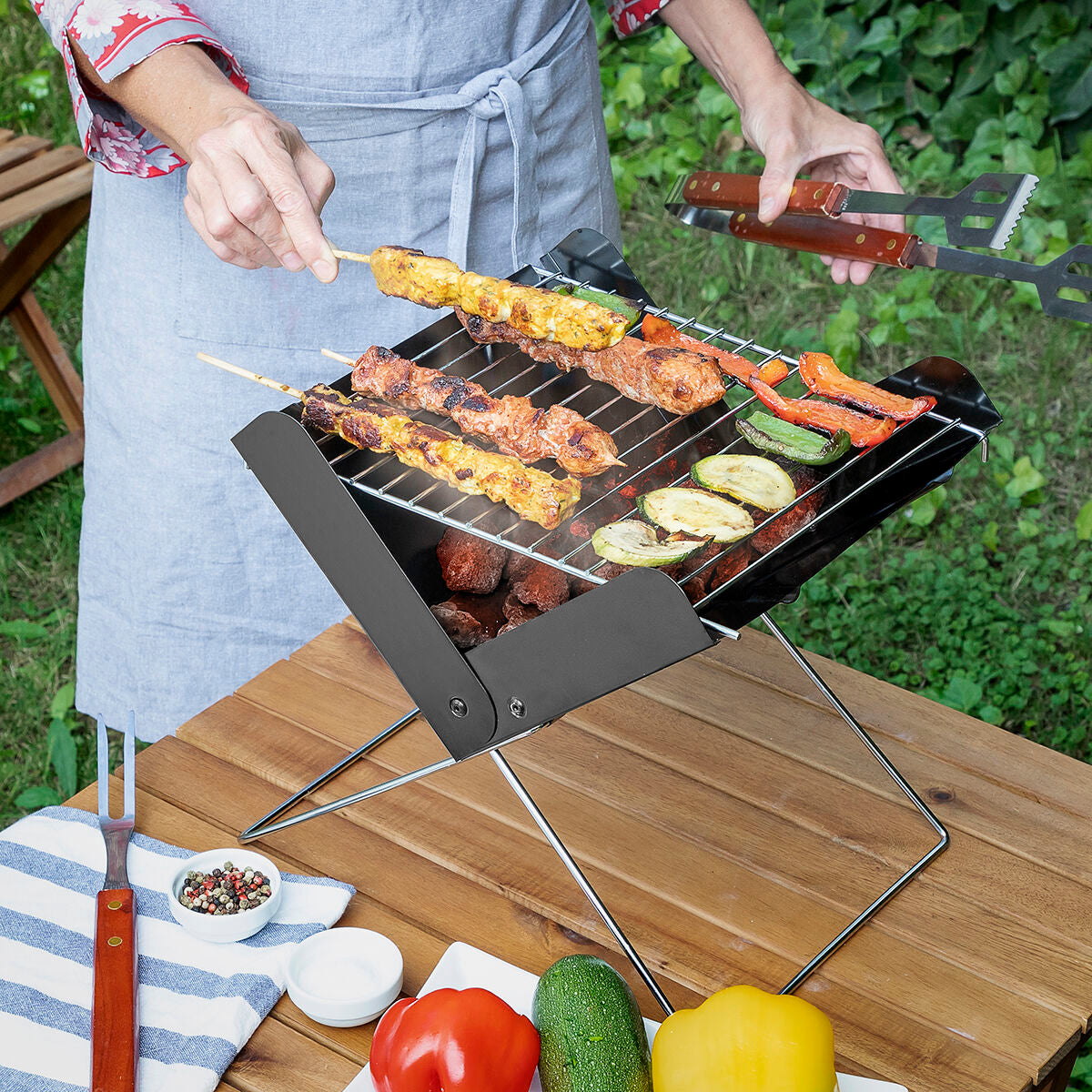A lifestyle image showing the barbecuing process using the Mini Folding Portable Barbecue. A person is grilling skewers and vegetables on a tabletop grill, set outdoors on a wooden table. The grill is filled with charcoal and food items like zucchini, peppers, and skewers of meat and vegetables.
