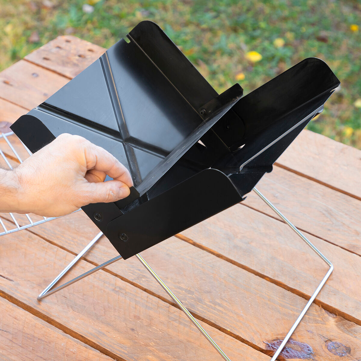 A person setting up the Mini Folding Portable Barbecue with the grill frame. It’s displayed outdoors on a wooden table.