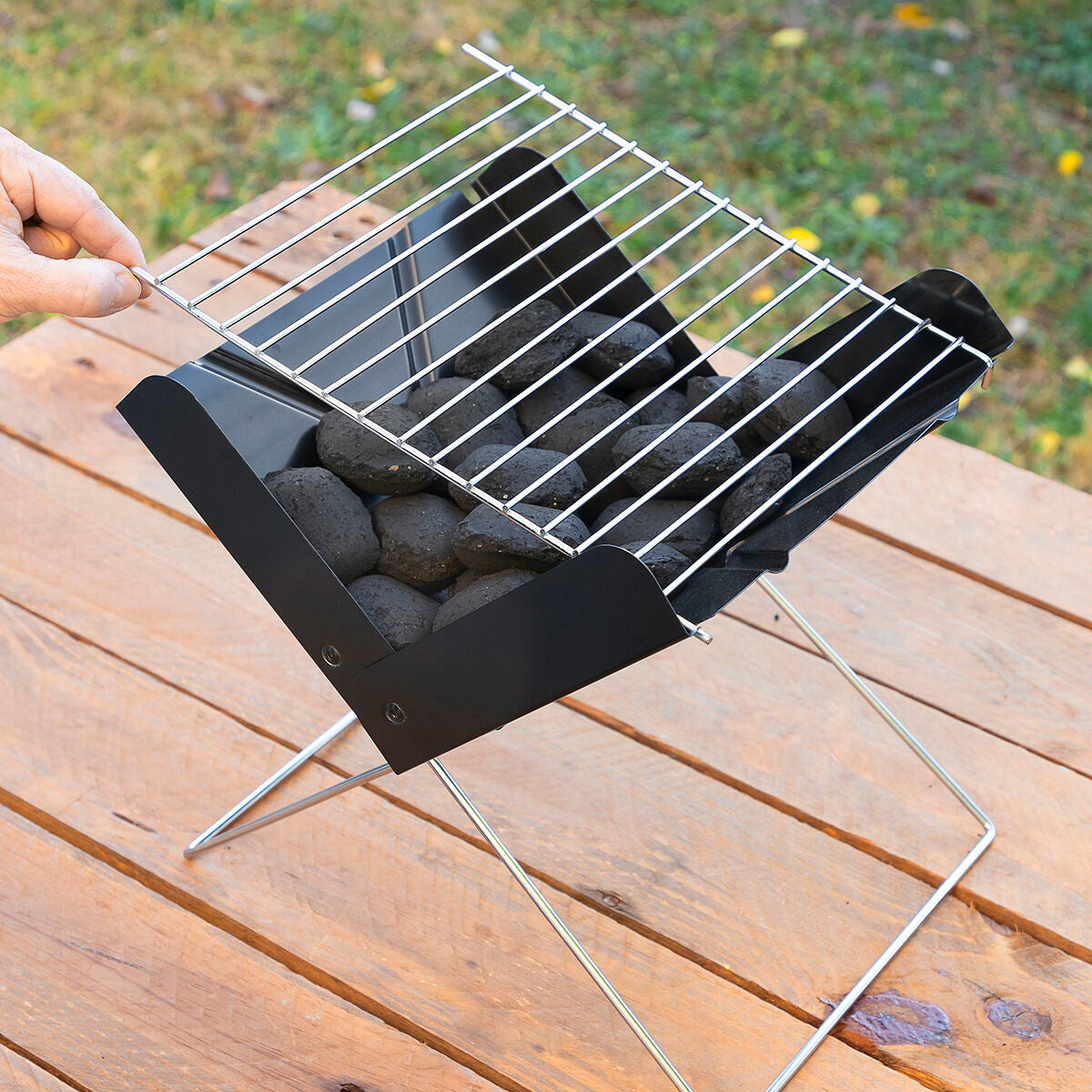 A close-up of the Mini Folding Portable Barbecue being set up with charcoal placed inside. The grill is shown on a wooden surface in an outdoor environment.