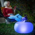 Young woman using the inflatable LED seat as a footrest while sitting on a chair outdoors at night. The seat is glowing blue, highlighting its LED feature. The setting is casual and relaxing, with greenery in the background.