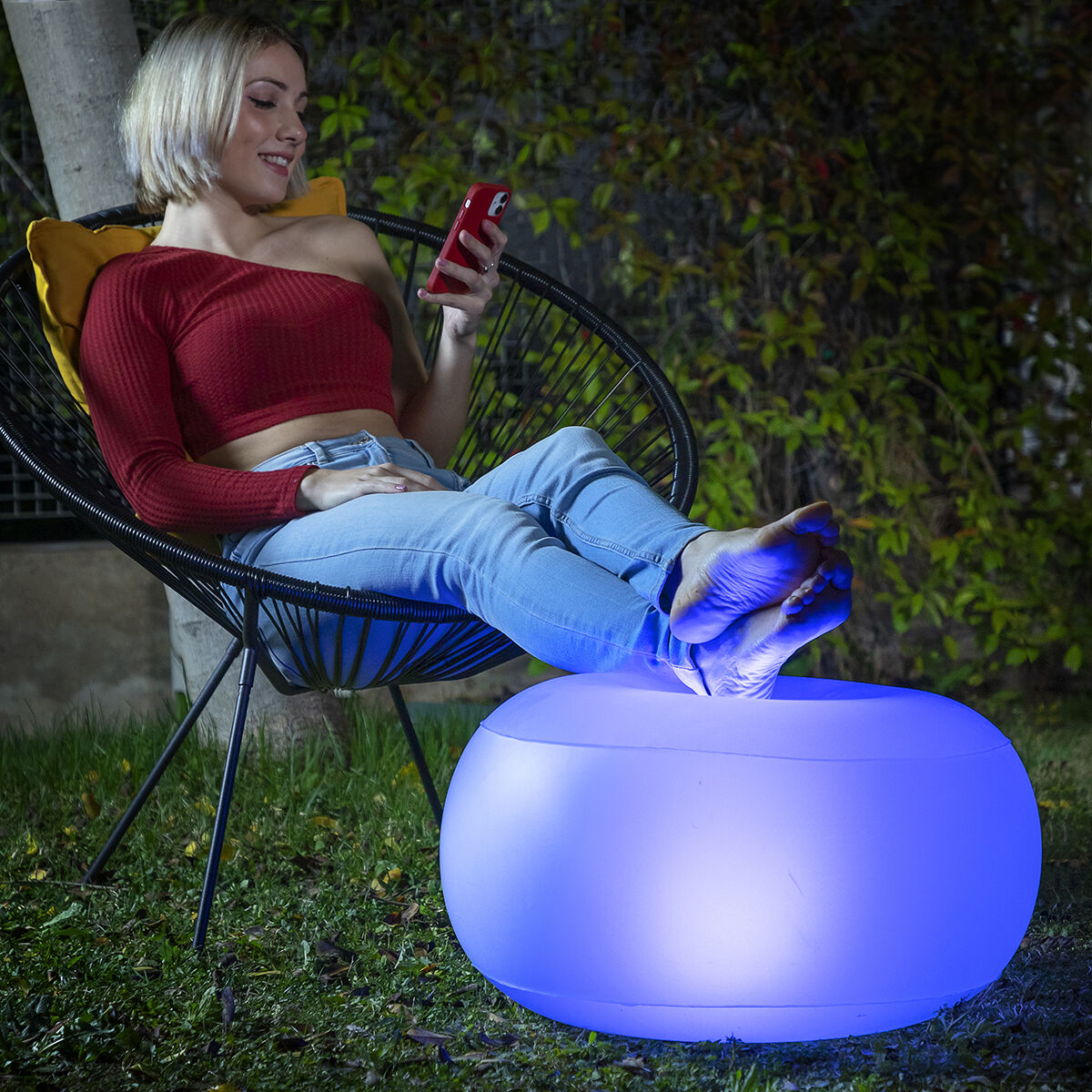 Young woman using the inflatable LED seat as a footrest while sitting on a chair outdoors at night. The seat is glowing blue, highlighting its LED feature. The setting is casual and relaxing, with greenery in the background.
