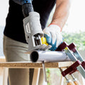 A person using the InnovaGoods Jigsaw Drill Attachment. The gray attachment is connected to a drill and is being used to cut a metal pipe. The setting is a workshop environment with tools visible in the background.