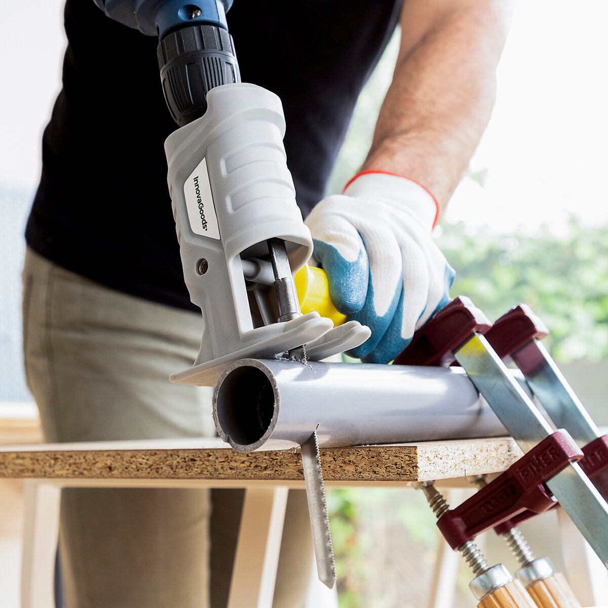 A person using the InnovaGoods Jigsaw Drill Attachment. The gray attachment is connected to a drill and is being used to cut a metal pipe. The setting is a workshop environment with tools visible in the background.