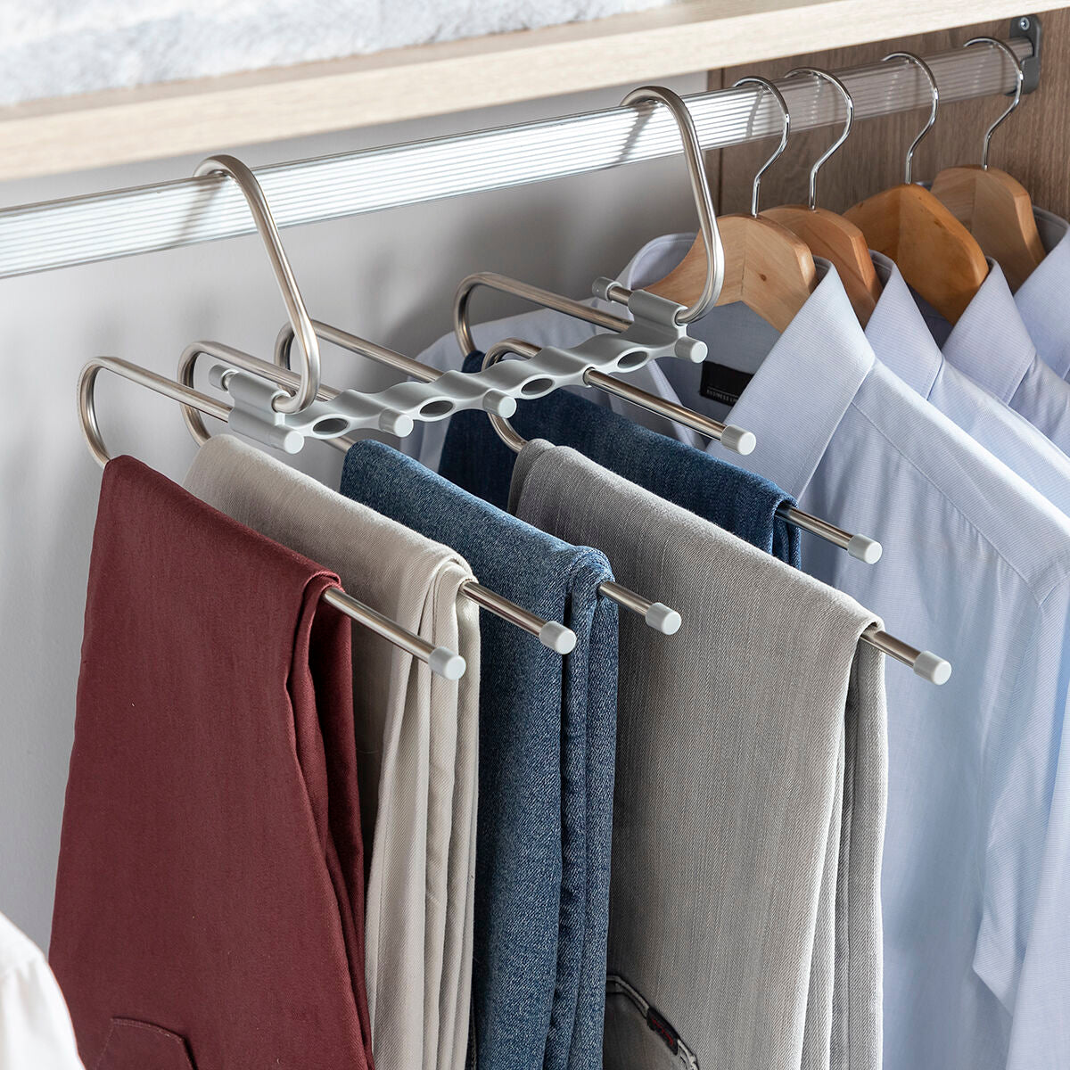 Hanger in use in a closet, holding various colored trousers with additional shirts on wooden hangers in the background.