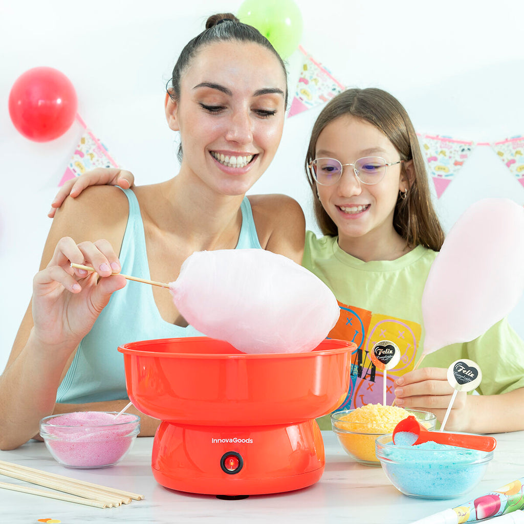 A lifestyle image showing a woman and a child making cotton candy using the Candy Floss Machine Cantty InnovaGoods. The machine is red and placed on a white table with colorful sugar bowls around. The setting is festive with balloons and decorations in the background.