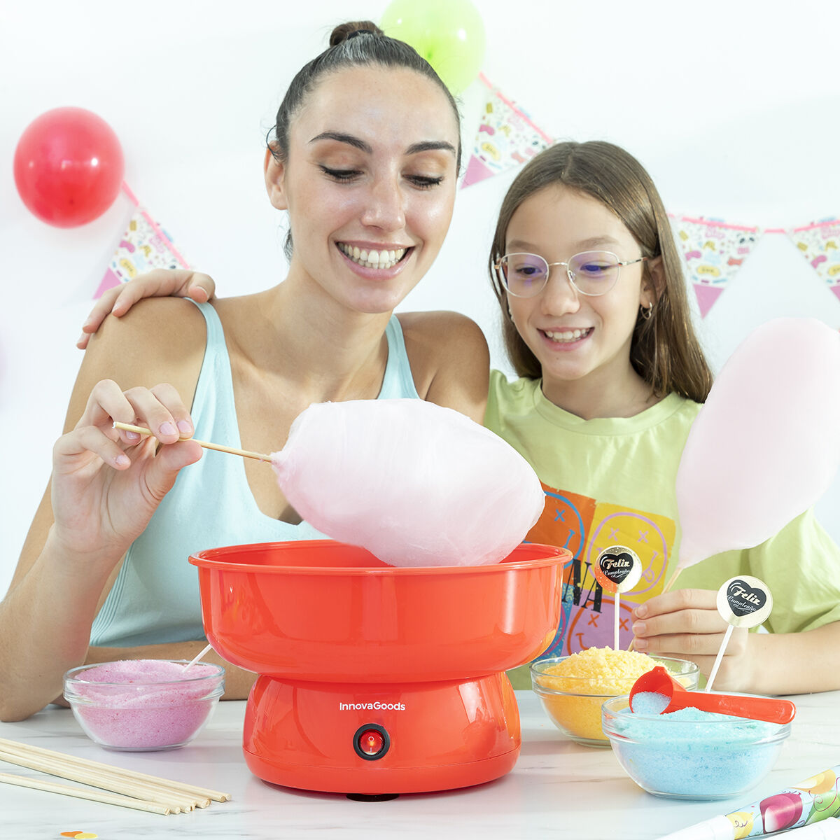 A lifestyle image showing a woman and a child making cotton candy using the Candy Floss Machine Cantty InnovaGoods. The machine is red and placed on a white table with colorful sugar bowls around. The setting is festive with balloons and decorations in the background.