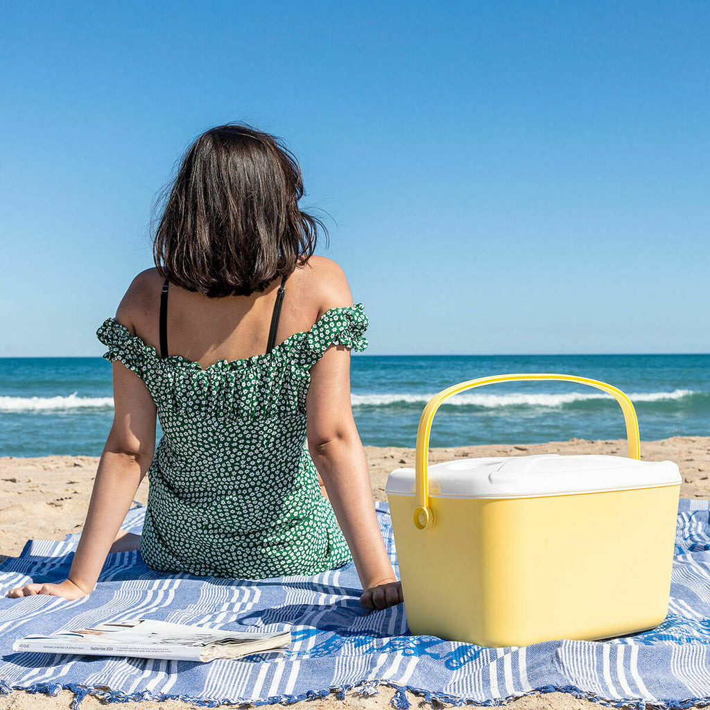 A yellow and white portable cooler being used on a beach. A person in a green dress is seated on a blue towel beside it, facing the ocean.