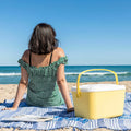 A yellow and white portable cooler being used on a beach. A person in a green dress is seated on a blue towel beside it, facing the ocean.