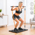 A woman is using the portable training system, performing a squat with the bar on her shoulders, attached to orange elastic bands on a black platform. The setting is indoors.
