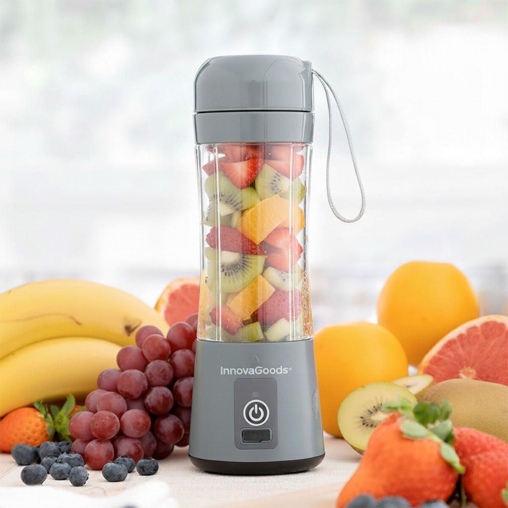 The blender is displayed filled with chopped fruits like strawberries, kiwi, and orange, emphasizing its use for fresh smoothies. It has a gray base, with the InnovaGoods logo and a power button. The background includes various whole fruits, creating a vibrant, healthy setting.