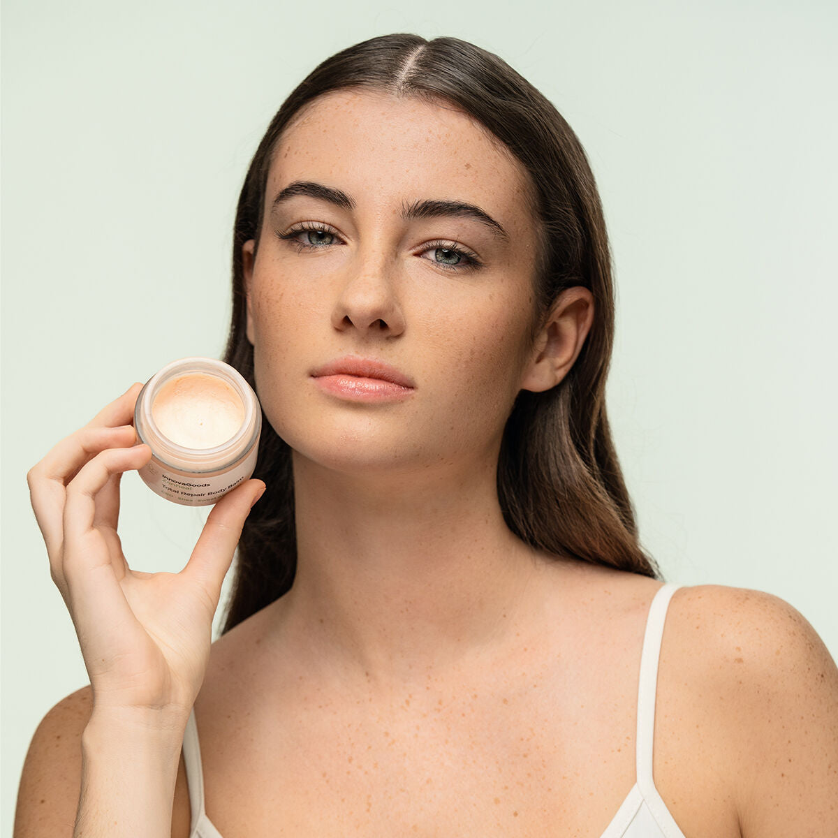 A close-up image of a person holding the open balm jar, showcasing the product's texture and rich consistency. The background remains soft green, highlighting the natural aspect of the product.