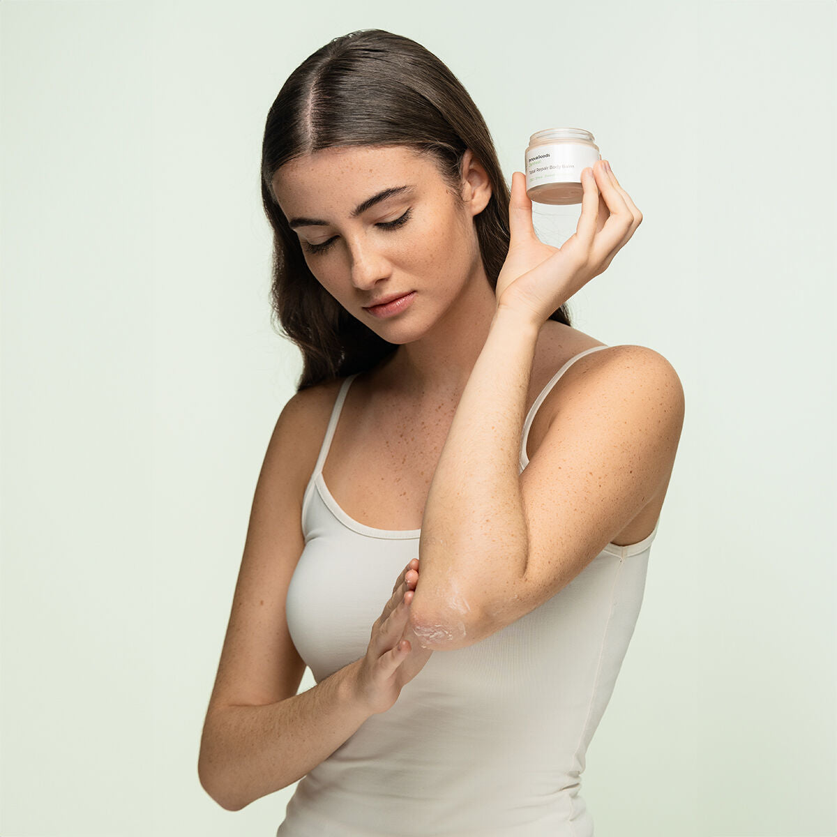 A person with long hair wearing a white tank top holds the balm jar while applying it to her arm. The setting is clean and minimalistic, adding a human element to the product usage.