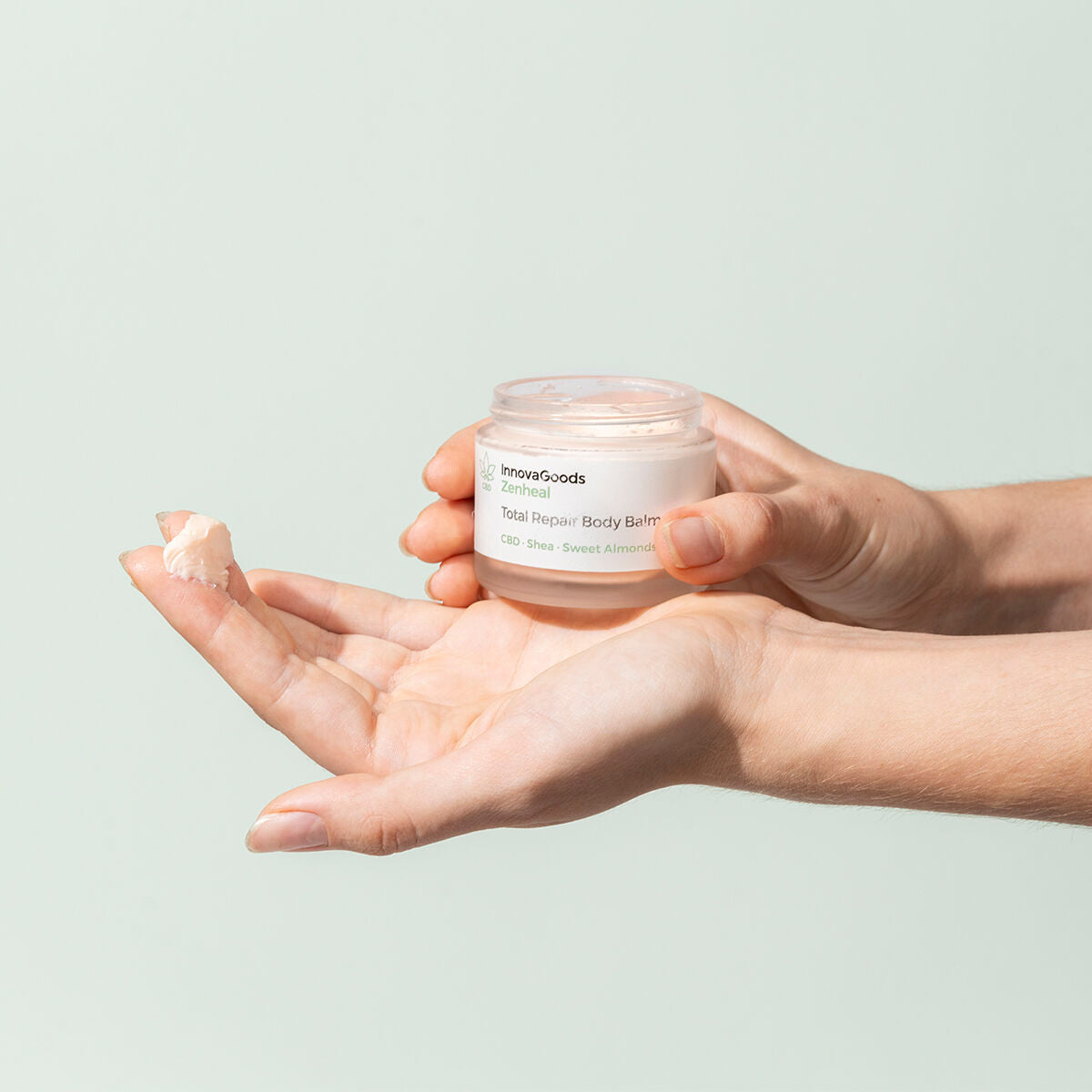 A close-up of hands holding the balm jar and applying a small amount to the skin. The background is a soft pastel green, emphasizing a gentle, nurturing application process.