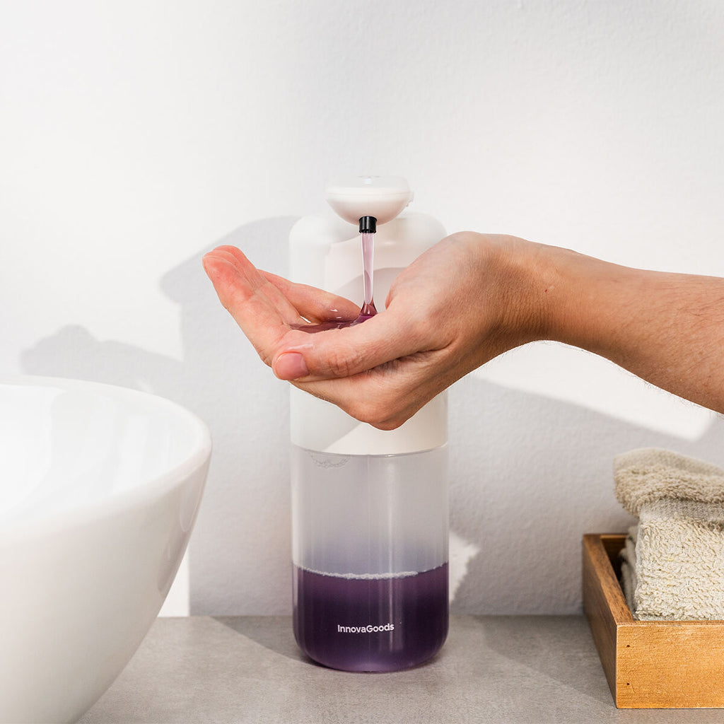 A hand using the dispenser, dispensing purple soap. The product has a white top and clear bottom, with the brand 'InnovaGoods' visible. It's situated on a bathroom counter next to a sink.