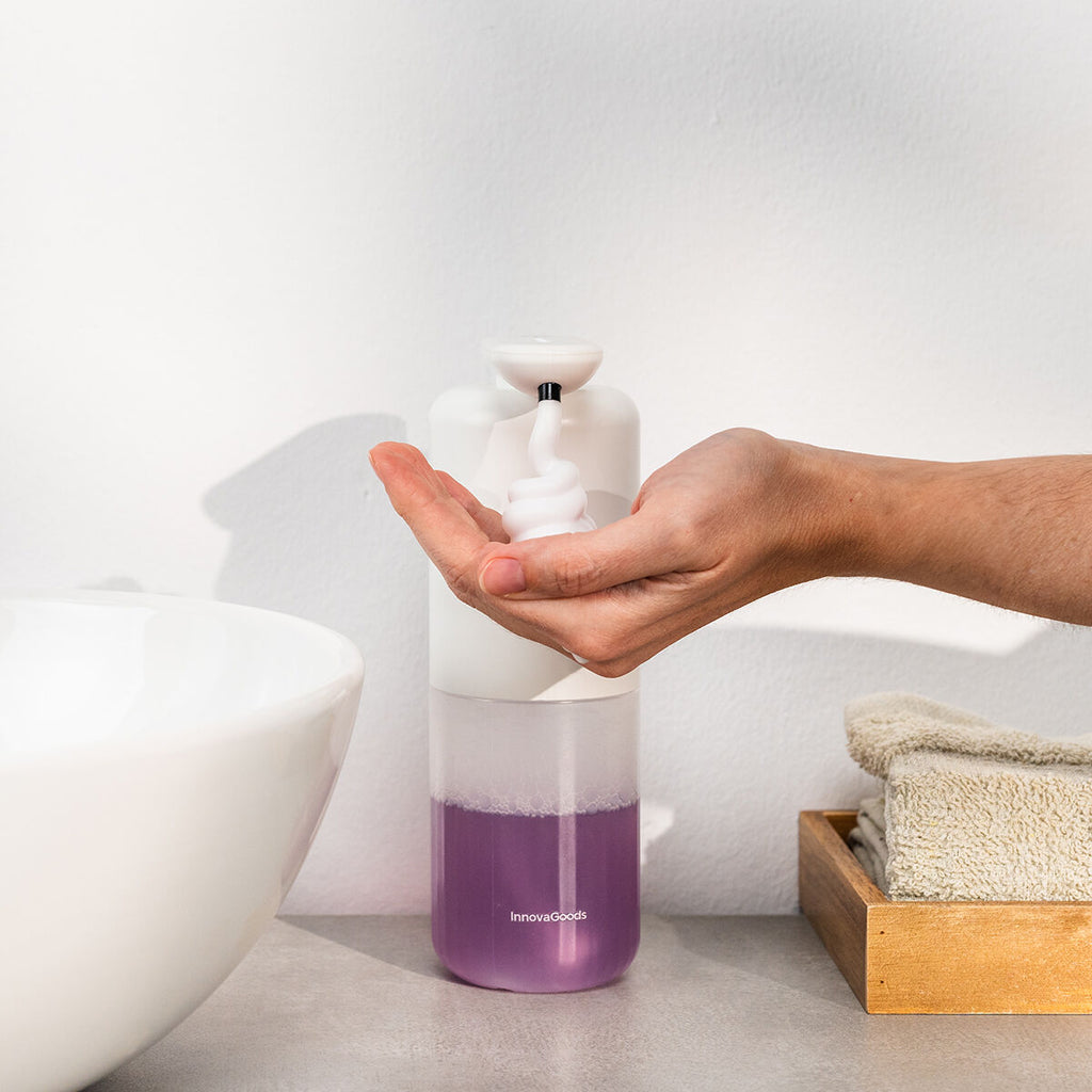 A hand is shown receiving foam from the white and transparent foaming soap dispenser, which is placed on a bathroom countertop next to a sink and towels. The soap in the dispenser is purple.