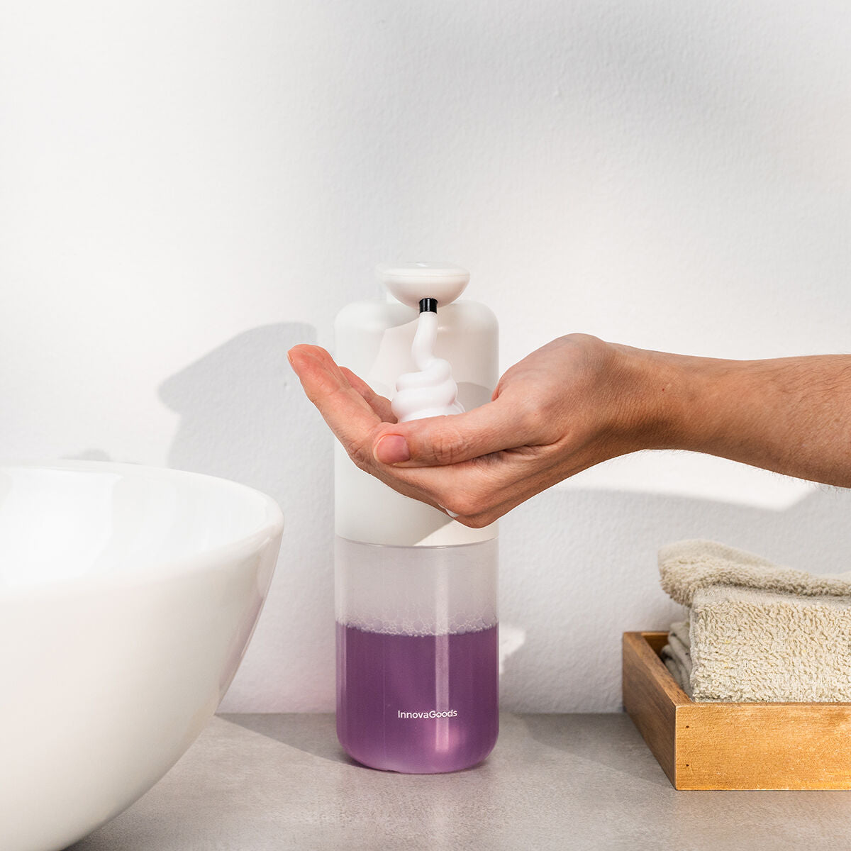 A hand is shown receiving foam from the white and transparent foaming soap dispenser, which is placed on a bathroom countertop next to a sink and towels. The soap in the dispenser is purple.
