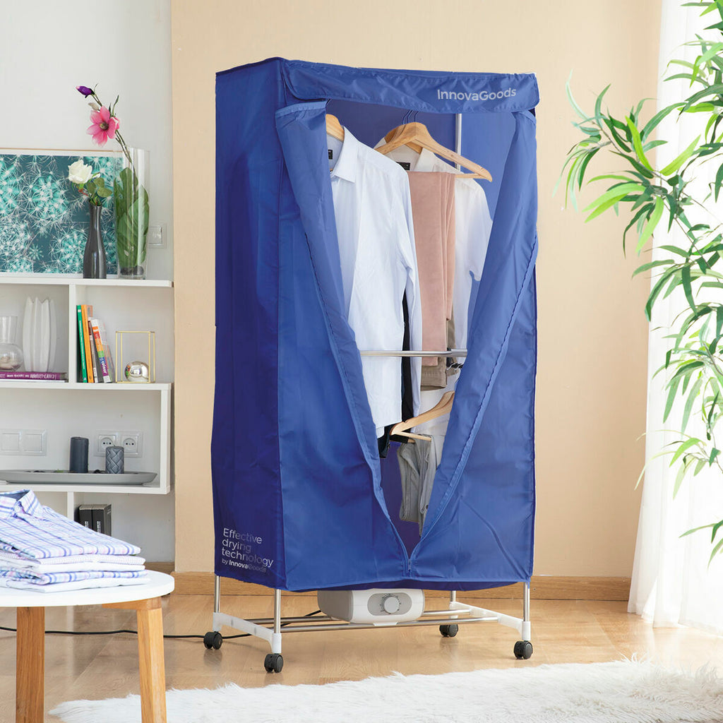 The dryer is assembled with a blue fabric cover open to reveal clothes hanging inside on metal rods. The setting is a living room corner with a wooden floor and a plant nearby. The front flap is open, and the dryer is standing on four small wheels for mobility.