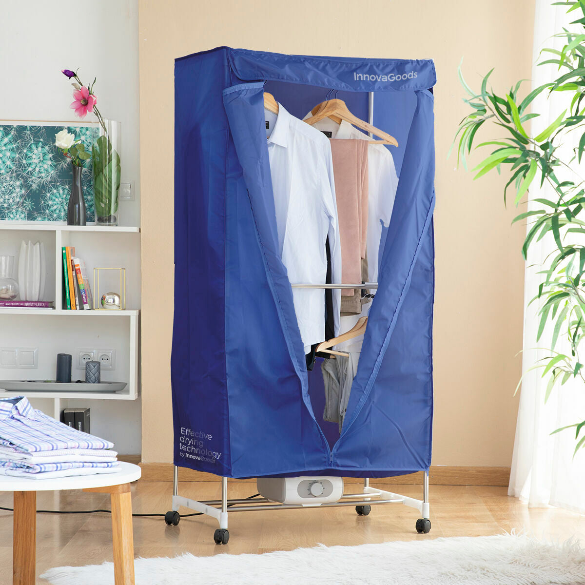 The dryer is assembled with a blue fabric cover open to reveal clothes hanging inside on metal rods. The setting is a living room corner with a wooden floor and a plant nearby. The front flap is open, and the dryer is standing on four small wheels for mobility.