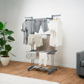A lifestyle image showing the clothes dryer in use, holding a variety of clothes such as shirts and trousers in a home setting with wooden floors and a potted plant nearby.