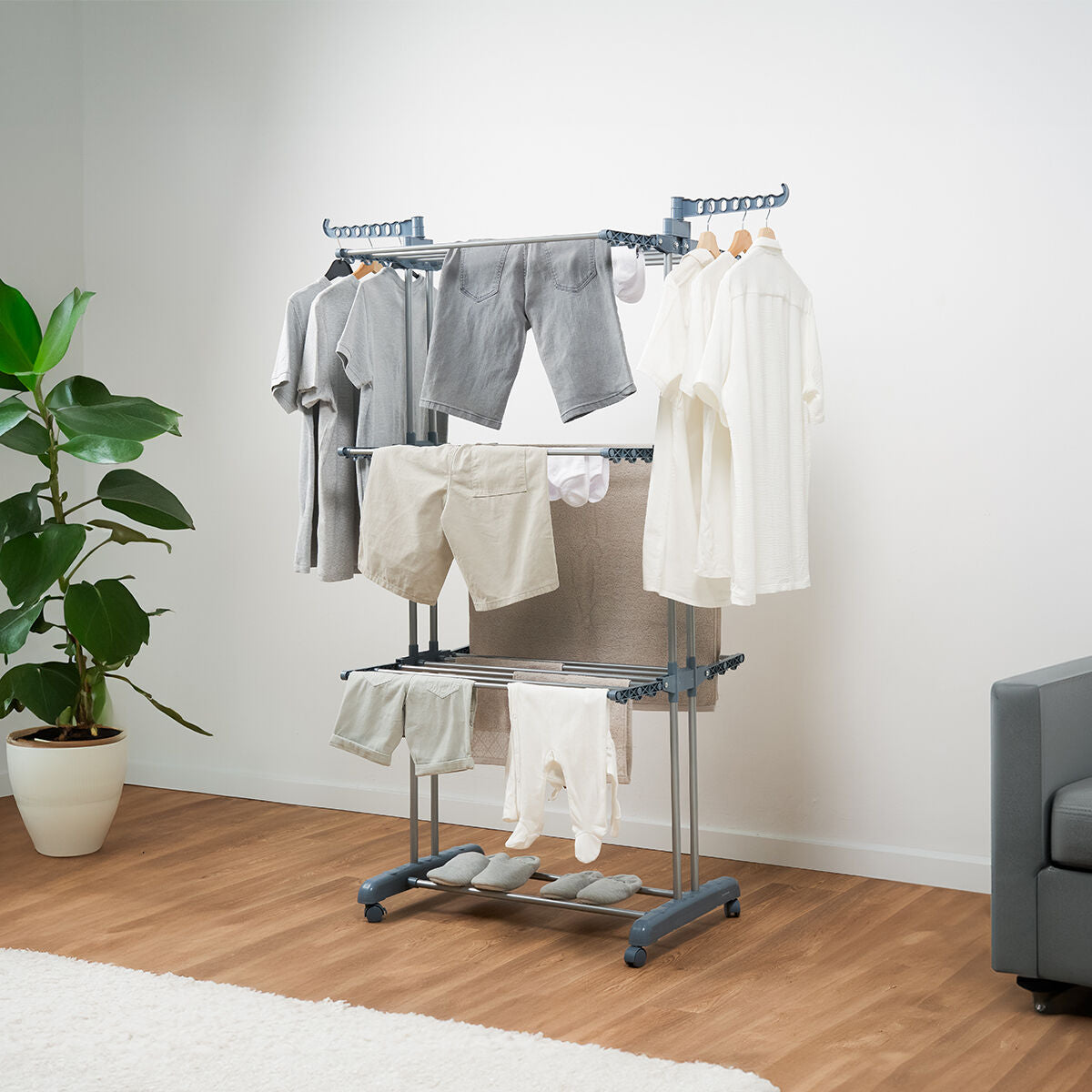 A lifestyle image showing the clothes dryer in use, holding a variety of clothes such as shirts and trousers in a home setting with wooden floors and a potted plant nearby.
