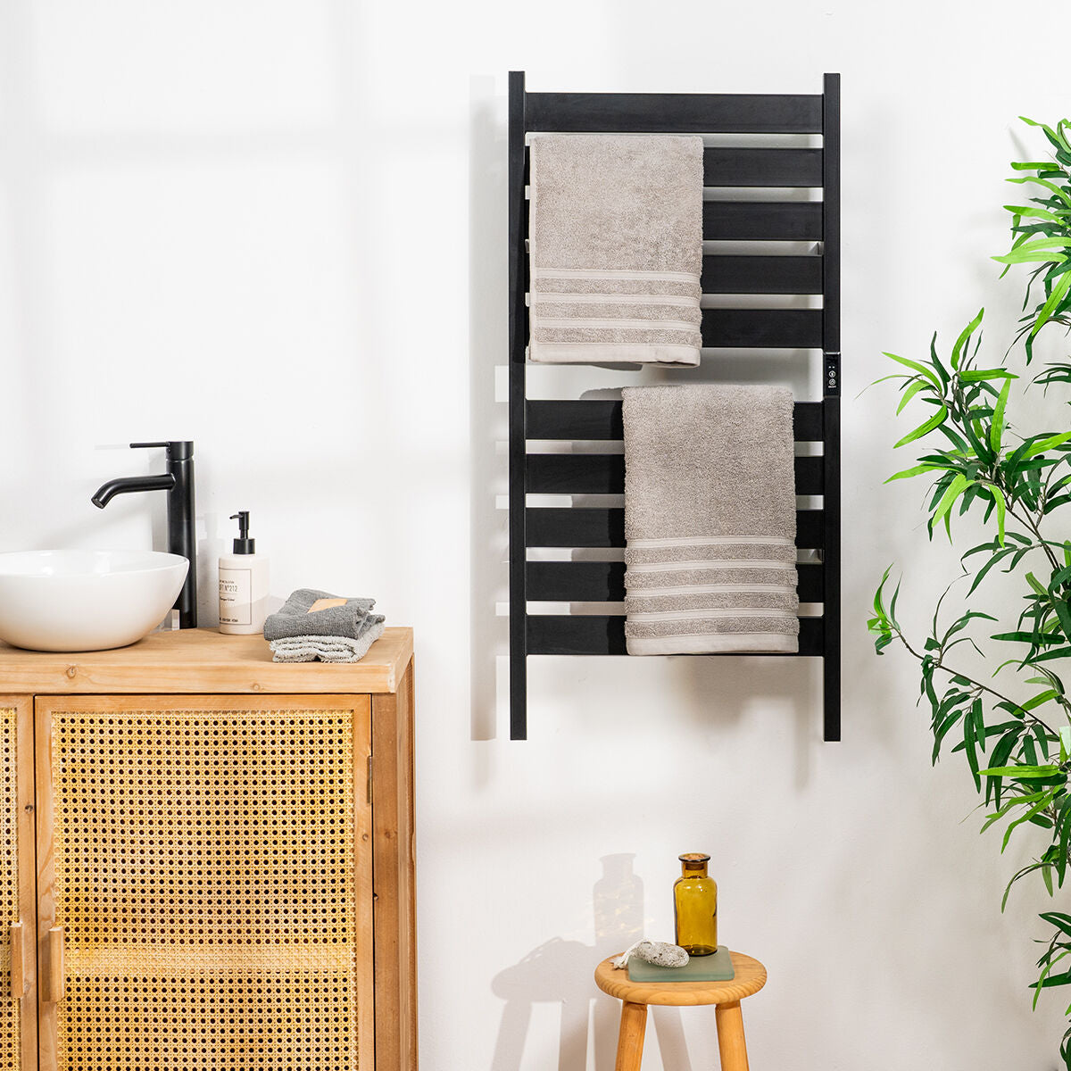 The towel rail is wall-mounted in a bathroom setting, alongside wooden and wicker furniture, with folded towels hanging. The aesthetic is clean and natural, with a plant adding a touch of greenery.