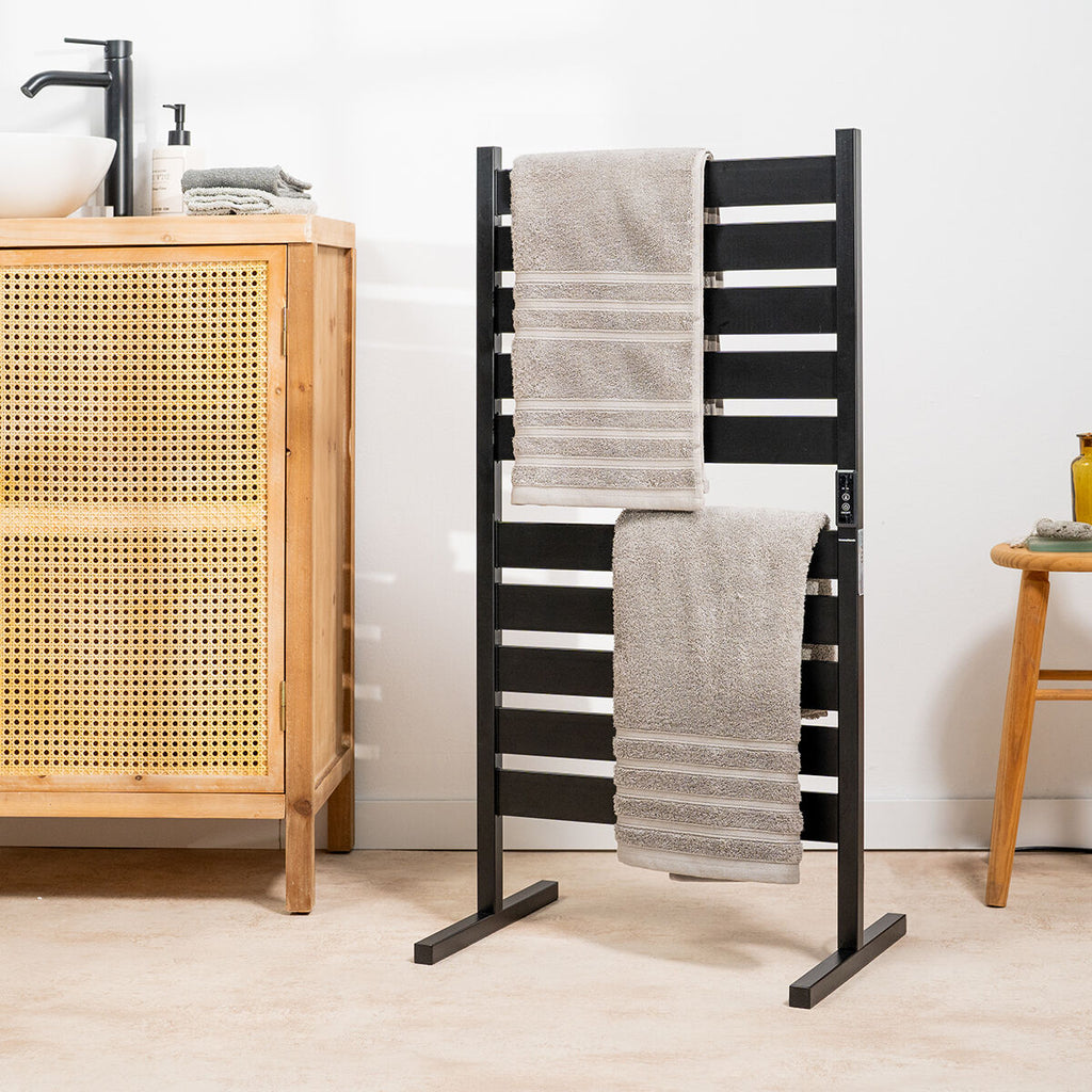 The towel rail positioned as a floor model in a bathroom, with towels draped and a blend of wooden and wicker furnishing around.