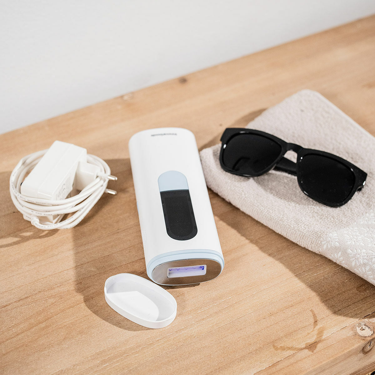 The Pulsed Light Hair Remover is placed on a wooden surface with its power adapter and sunglasses nearby, showcasing the included accessories.