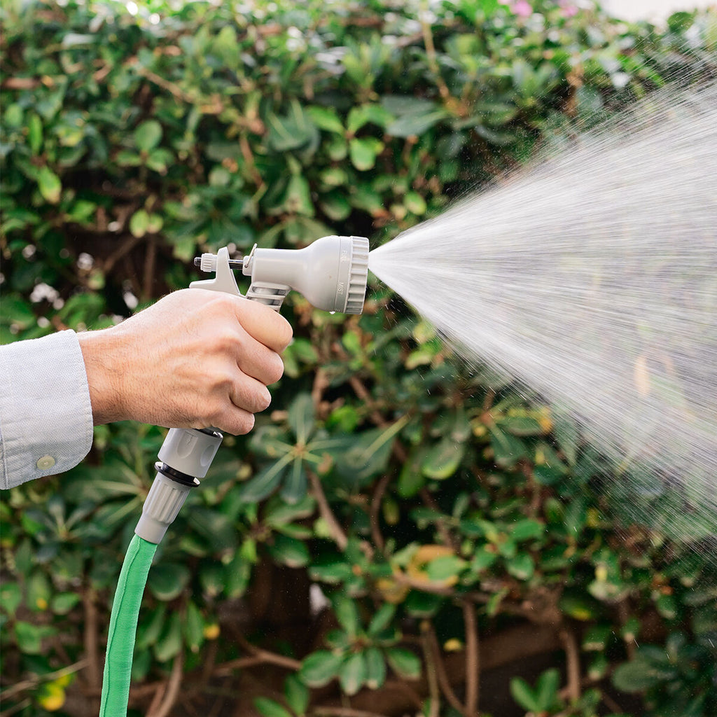 Close-up of a hand holding the grey spray nozzle of the green expandable hose, showing the nozzle spraying water in a mist over a leafy background.