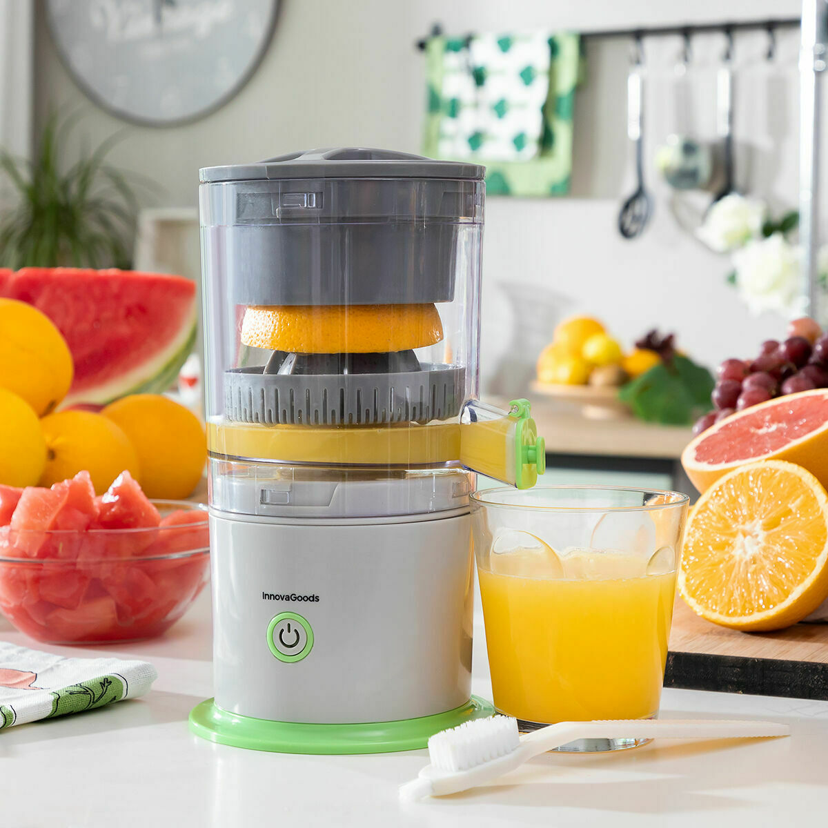 The juicer in a kitchen setting with oranges and a toothbrush nearby. Glass with orange juice is placed to the side.