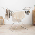 The image displays the Foldable Electric Clothes Dryer in an indoor setting. It is holding several towels and clothes, emphasizing its functionality. The dryer is silver, with two extended wings and a sturdy frame. The setting includes a chair and a cabinet, giving a cozy room vibe.