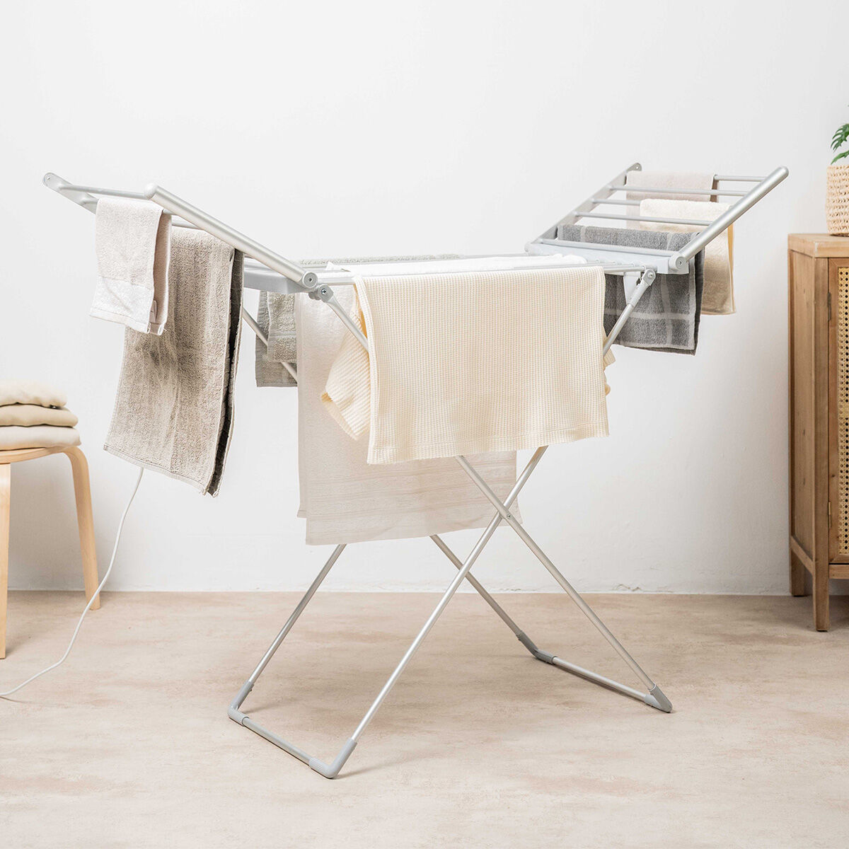 The image displays the Foldable Electric Clothes Dryer in an indoor setting. It is holding several towels and clothes, emphasizing its functionality. The dryer is silver, with two extended wings and a sturdy frame. The setting includes a chair and a cabinet, giving a cozy room vibe.