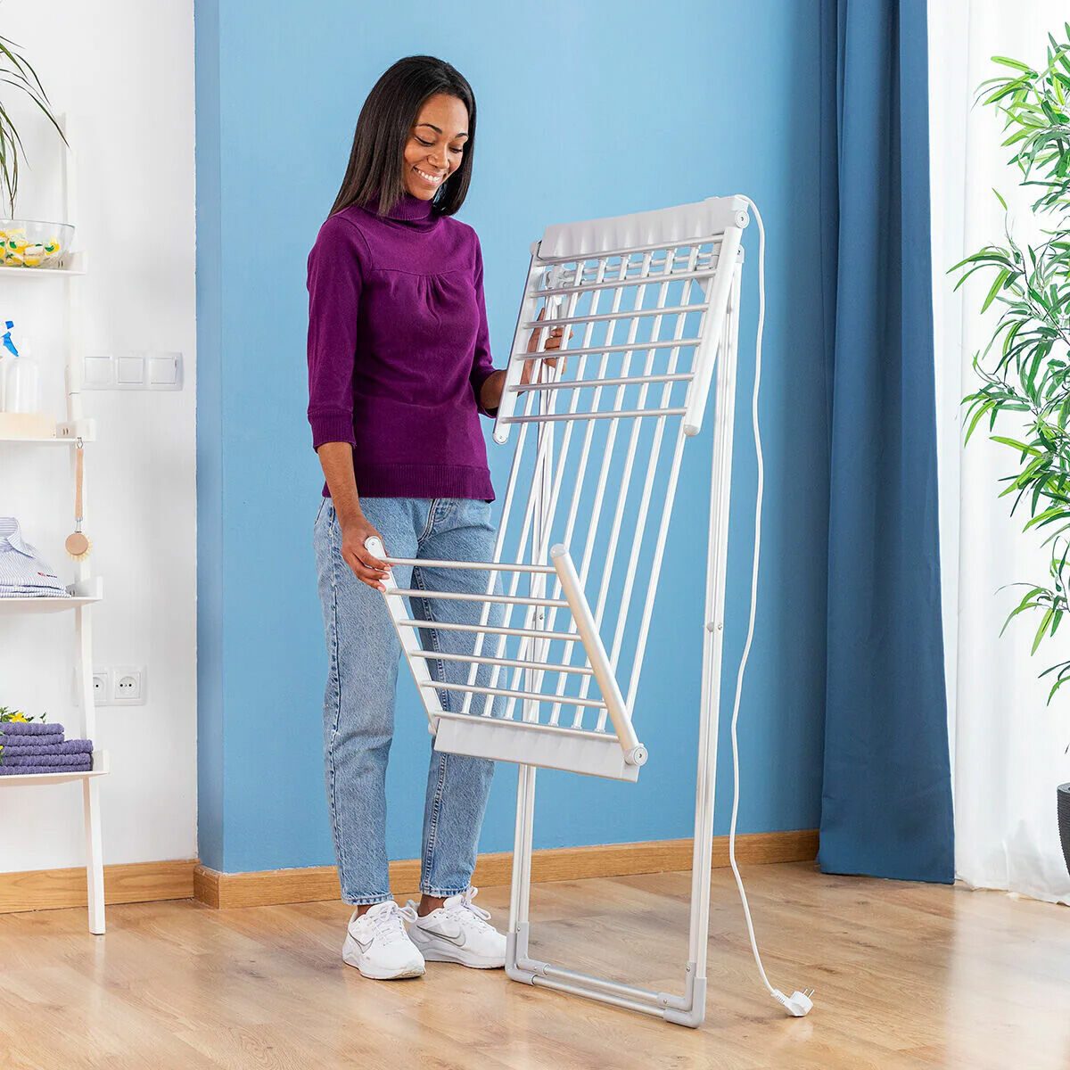 A person is seen unfolding the dryer, illustrating its ease of setup. The setting is a modern home interior with blue walls.