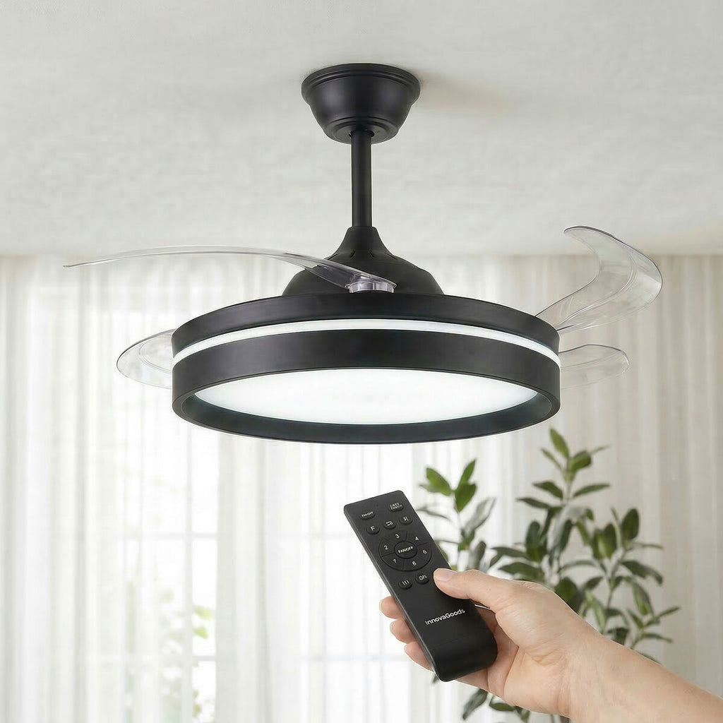 A modern black ceiling fan with retractable transparent blades and integrated LED light. A hand holding a remote control is visible, suggesting ease of use. The setting is a well-lit room with plants in the background.