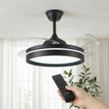 A modern black ceiling fan with retractable transparent blades and integrated LED light. A hand holding a remote control is visible, suggesting ease of use. The setting is a well-lit room with plants in the background.