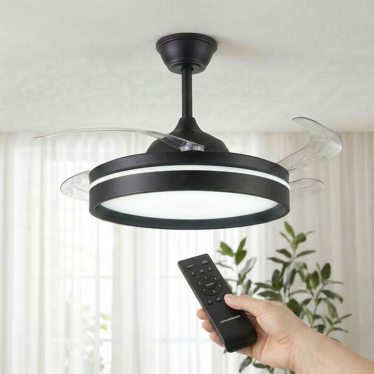 A modern black ceiling fan with retractable transparent blades and integrated LED light. A hand holding a remote control is visible, suggesting ease of use. The setting is a well-lit room with plants in the background.