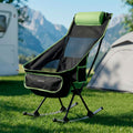 The chair is set up outdoors on grass, with a camper and tent in the background. It is black with green accents, including a cushion for the head.