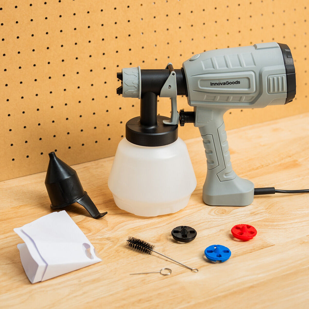 The paint sprayer gun is displayed disassembled on a wooden surface, including a gray and black electric paint sprayer with a white paint container, a black funnel, a brush, and three detachable spray nozzles in black, red, and blue.