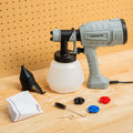 The paint sprayer gun is displayed disassembled on a wooden surface, including a gray and black electric paint sprayer with a white paint container, a black funnel, a brush, and three detachable spray nozzles in black, red, and blue.
